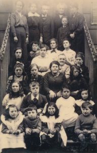 A photo of children and adults on the front steps of a building. There are some children sat at the front. The children have lots of different expressions. Some of the children are smiling, some look distracted, most of them have neutral expressions. There are some adults standing at the back of the steps. It is harder to see their faces. This is because the photo has faded at the top from age. 