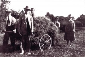There are 3 young men pushing a wagon full of hay. They are smiling and squinting because of the sun. They are wearing shirts, suspenders and ties. Two of them are wearing caps. One man is standing in front. He is wearing a suit and a fedora. An elderly Grace stands next to them in the field. She is wearing a long coat, a wide-brimmed felt sun hat, a high collar, and wellies. It is hard to see her face but it looks like she is slightly smiling.