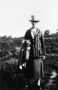 An elderly Grace is standing in a sunny field. She is wearing a stripy jacket, a three quarter-length skirt, and a big hat. She has what looks like a coat and scarf draped over one arm. She is smiling.